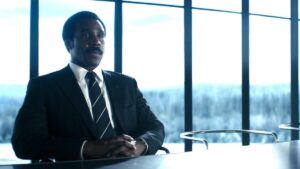 Seth Milchick in a suit sitting at a table with a snowy background behind the windows on Severance