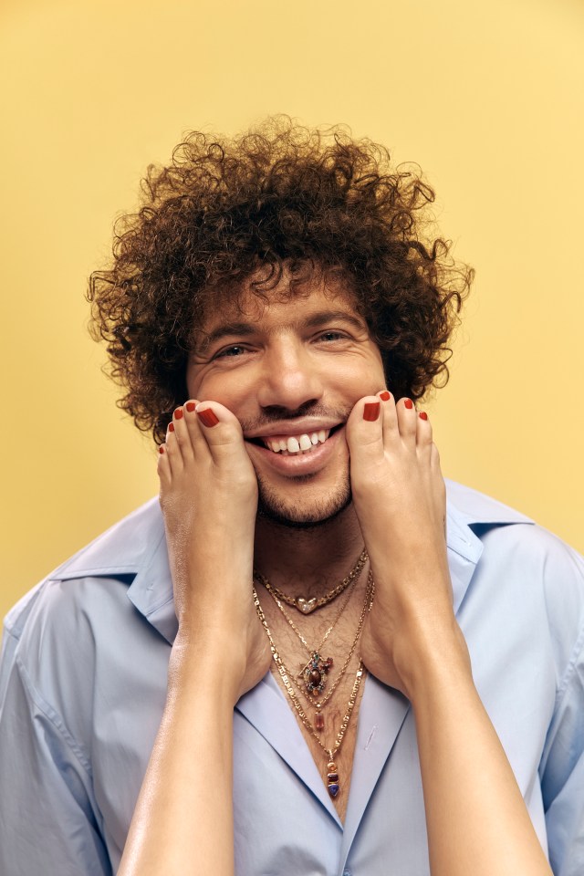 A man with curly hair smiles as a pair of feet hold his cheeks.