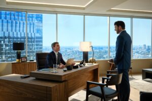 A man in a suit sits at a desk staring up at another man in a suit