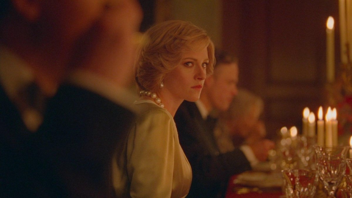 Kristen Stewart as Princess Diana in Spencer. Sitting on a long dining table packed with people only she is in focus.