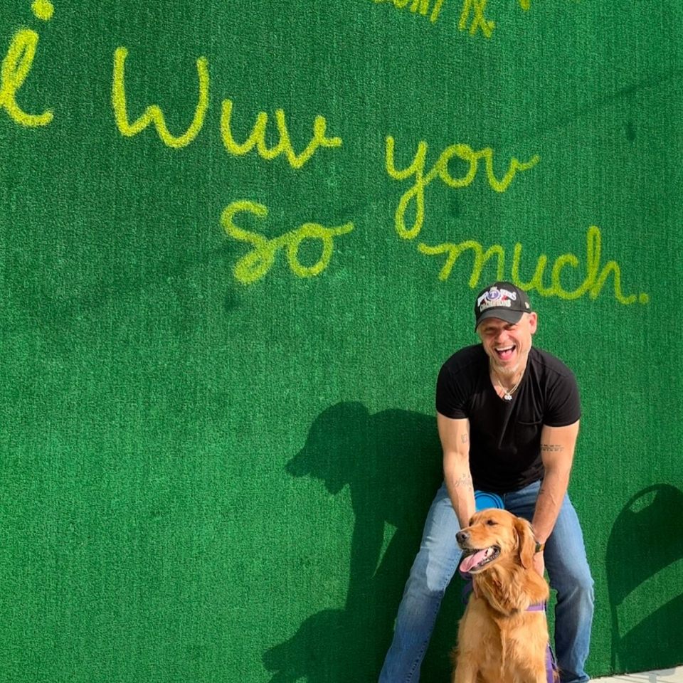 Aaron Schwartz, former child star, sits with his dog in front of a green wall with "I love you so much" written on it.