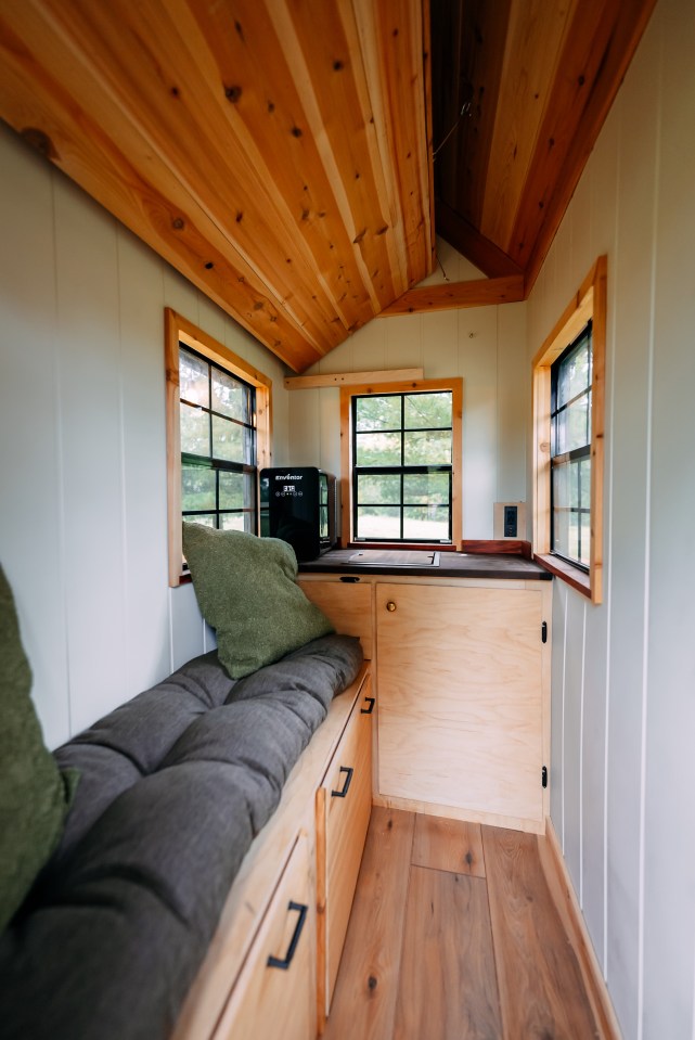 Interior of a tiny house with wood walls and floors, a built-in bench seat with cushions, and small windows.