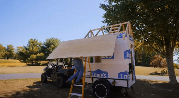 Person lifting a large piece of plywood onto a tiny house under construction.