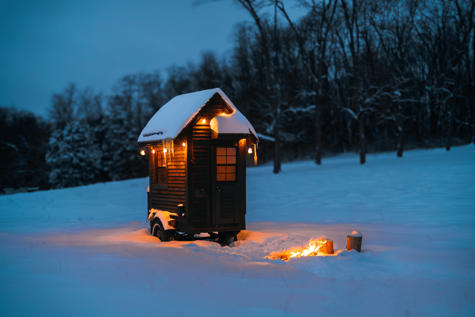 A snow-covered tiny house with lights on, sitting in a snowy field at night, with a small fire burning nearby.