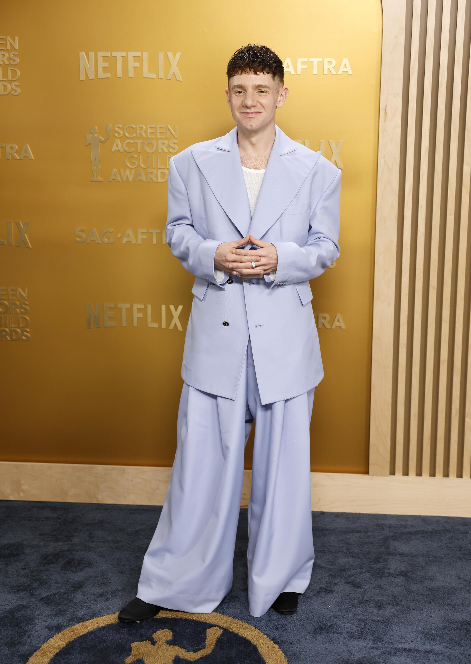 Chris Perfetti, in a purple oversized suit, smiles stands with hands clasped. 