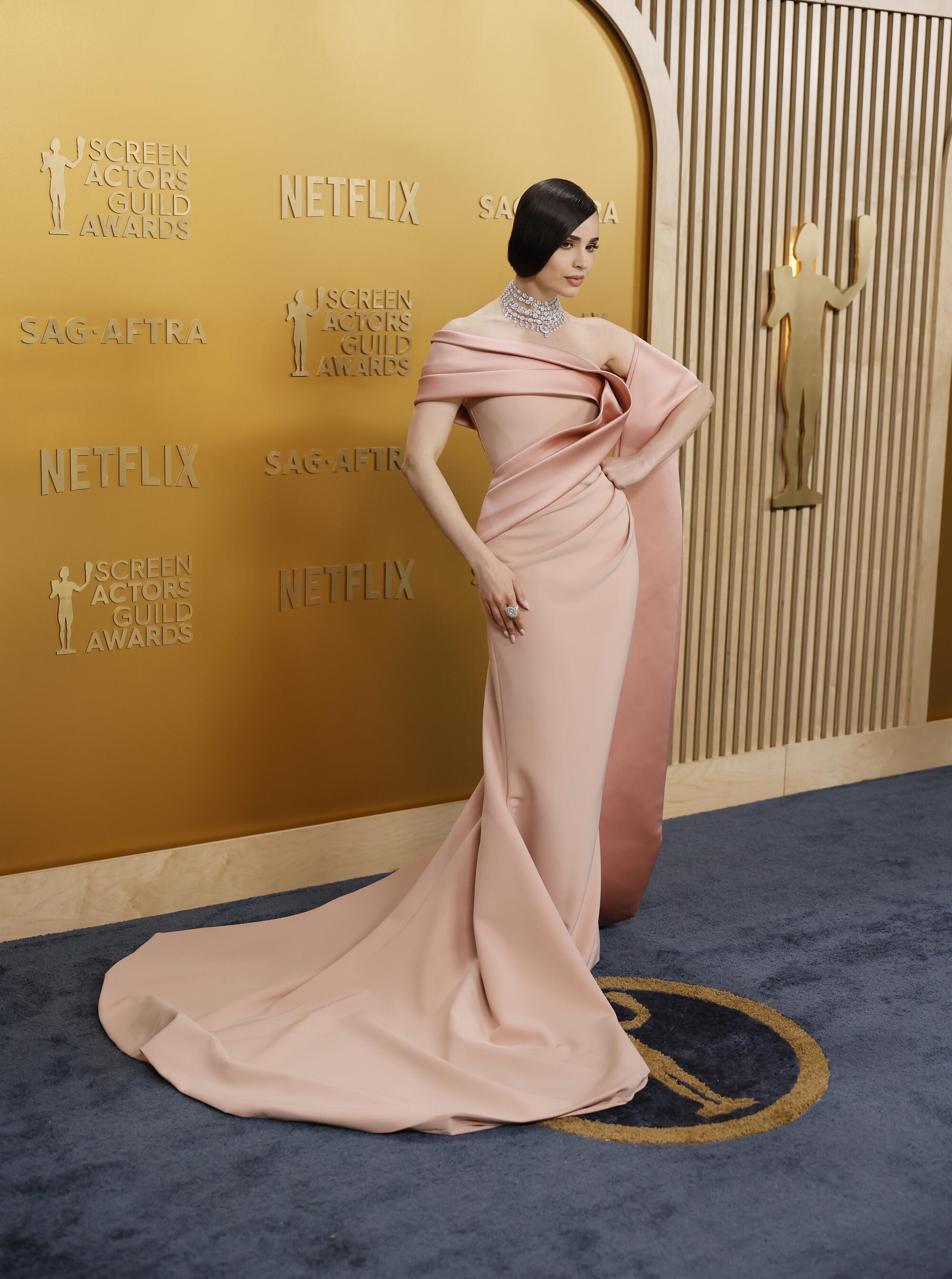 Sofia Carson, in a blush off the shoulder gown, poses with her fist on her hip.