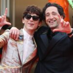 A photo of two men, Timothée Chalamet and Adrien Brody, smiling and posing for photographers with one arm around each other.