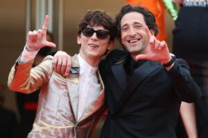 A photo of two men, Timothée Chalamet and Adrien Brody, smiling and posing for photographers with one arm around each other.