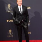 Conan O'Brien, in a black suit, stands with his hands in his pockets on a red carpet.