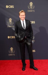 Conan O'Brien, in a black suit, stands with his hands in his pockets on a red carpet.