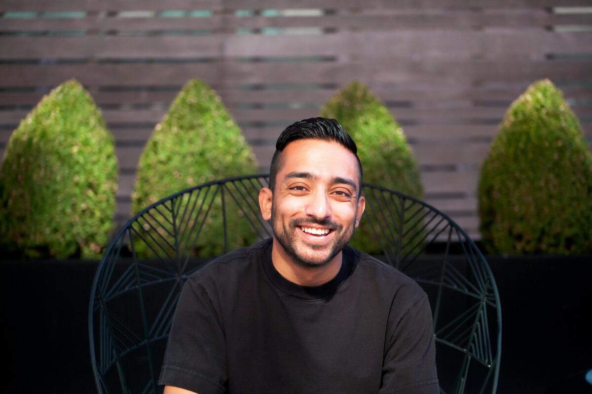 Mayukh Sen, in a black T-shirt, smiles and sits on a chair in front of hedges.