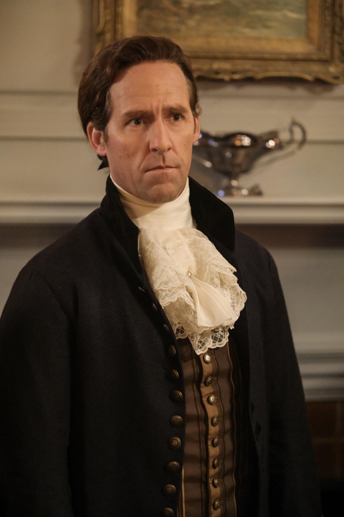 A man in a dark jacket, vest and ruffled collar standing with his back to a fireplace.