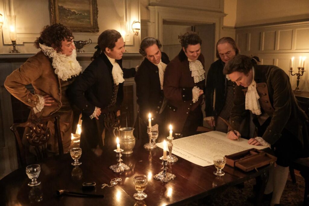 Six men standing around a candlelit table looking at a large document that is being signed.