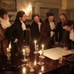 Six men standing around a candlelit table looking at a large document that is being signed.