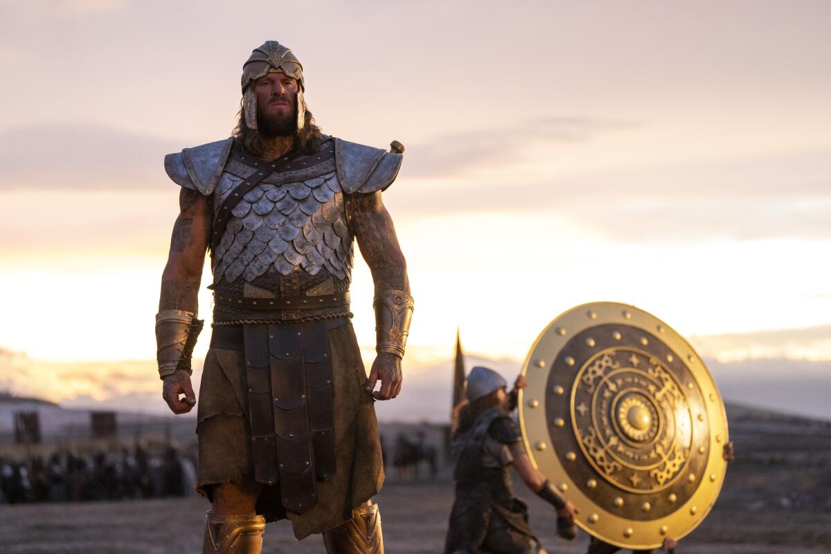 A giant man in metal and leather armor standing in a field as two smaller people carry away a golden shield.