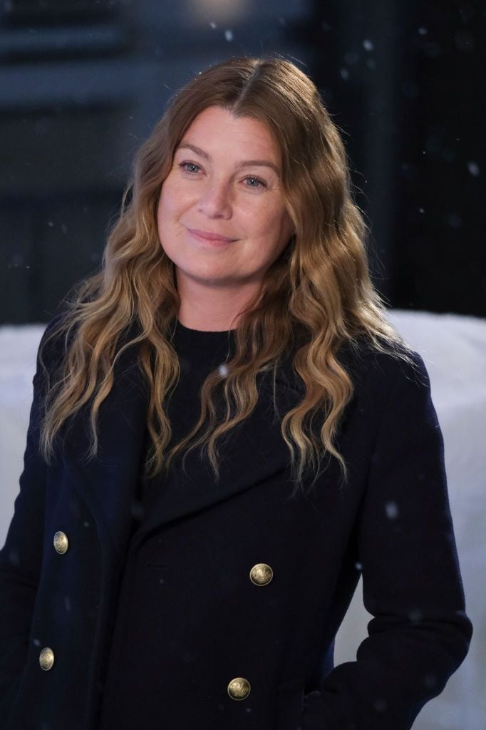 Ellen Pompeo in a navy coat with gold buttons, standing outdoors in a snowy setting, smiling gently.