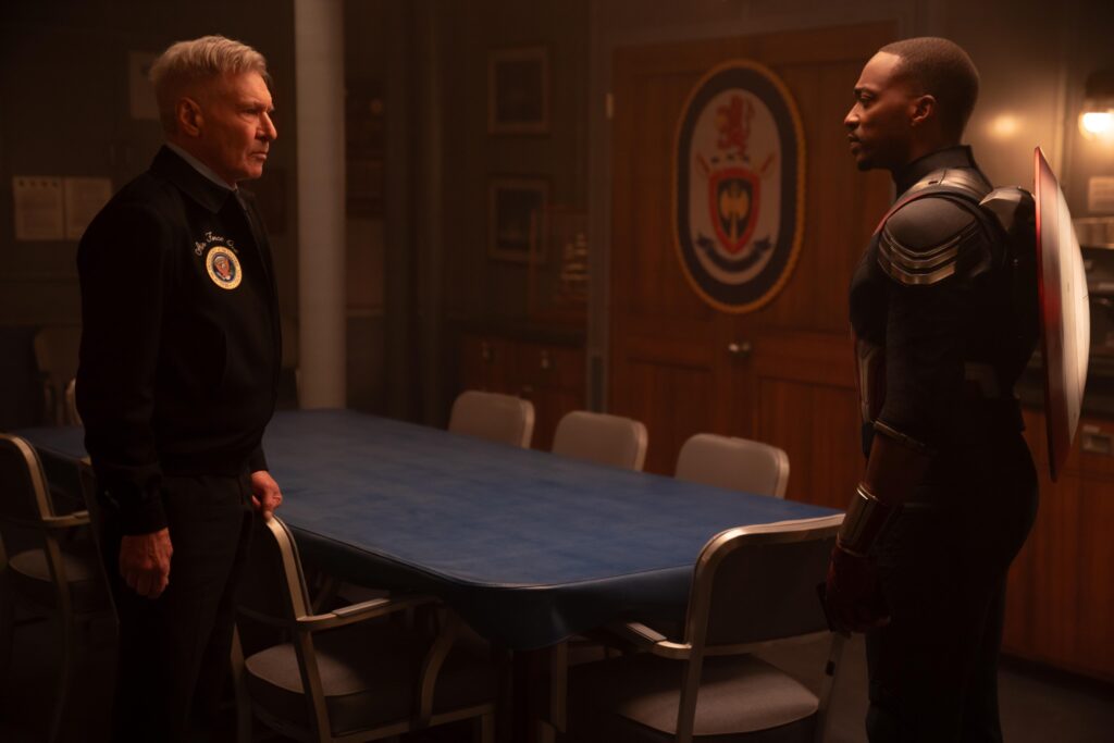 (L-R): Harrison Ford as President Thaddeus Ross and Anthony Mackie as Sam Wilson/Captain America Captain America: Brave New World. They’re looking at each other tensely in a room with a conference table and a military seal on the wall.