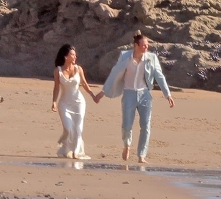 Kim Kardashian and Matthew Noszka on a beach, holding hands, in wedding attire.