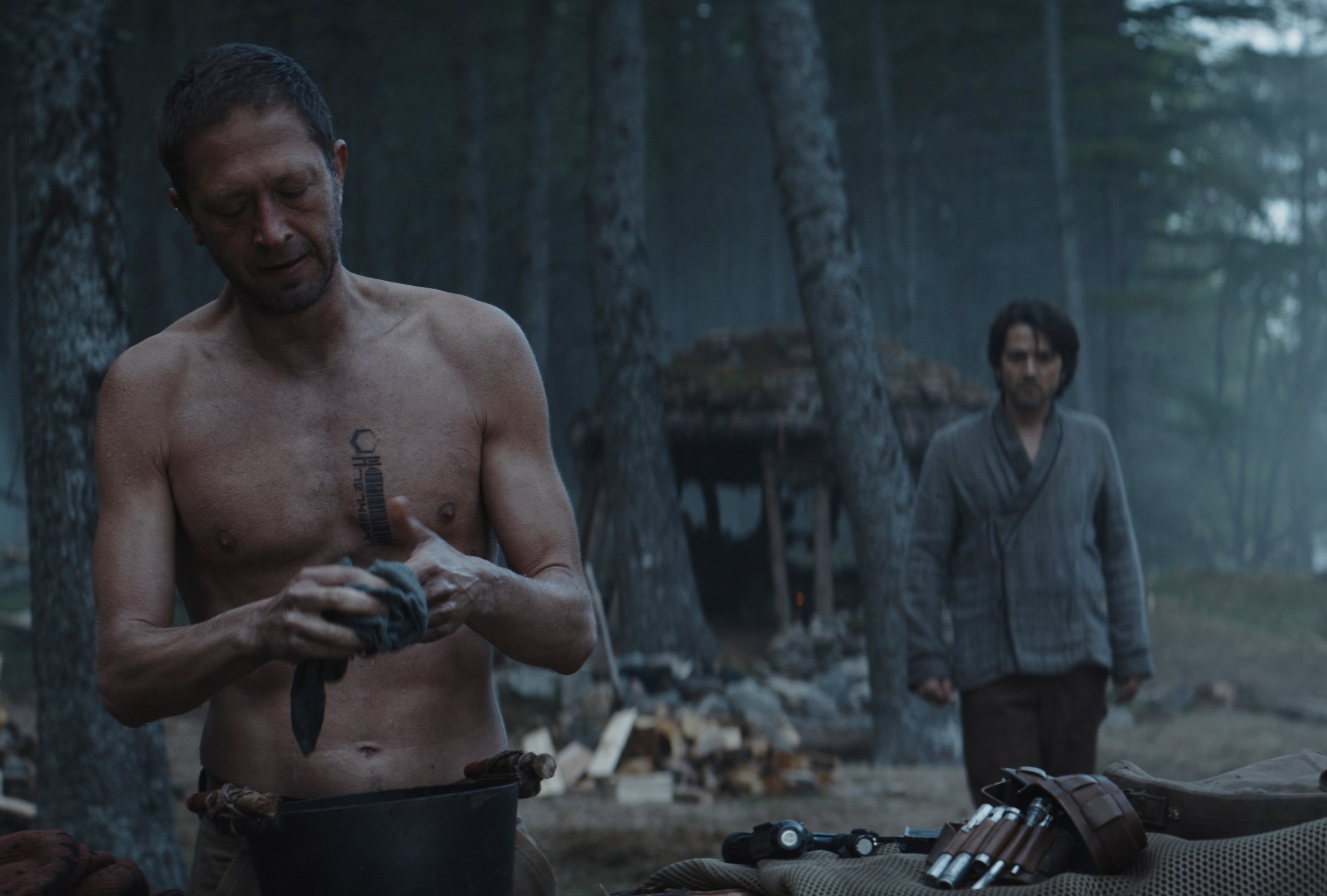 Seen stands shirtless, tinkering with a device in the Rebel camp while Cassian Andor walks towards him from behind