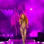 Shakira performs on stage in a patterned bodysuit, singing with a wolf graphic illuminated behind her in a purple-lit setting during her Las Mujeres Ya No Lloran tour.