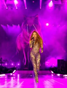 Shakira performs on stage in a patterned bodysuit, singing with a wolf graphic illuminated behind her in a purple-lit setting during her Las Mujeres Ya No Lloran tour.