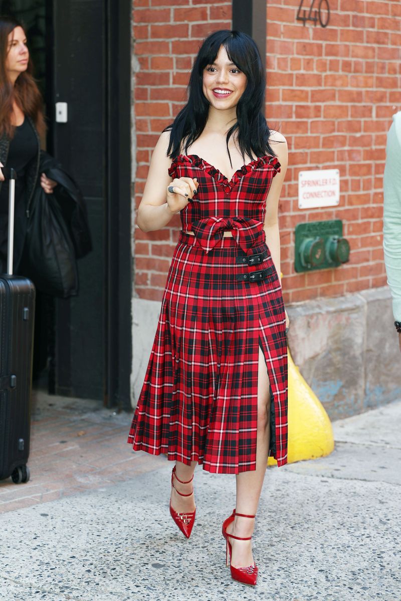 Jenna Ortega flashes a smile and waves as she steps out in New York City. The 21-year-old American actress strikes a fashionable pose in a red and black plaid dress paired with bright red heels.