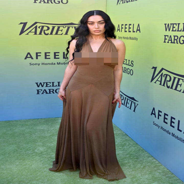 A person on the red carpet wearing a flowing, sleeveless gown with a deep neckline, posing in front of a backdrop with sponsor logos