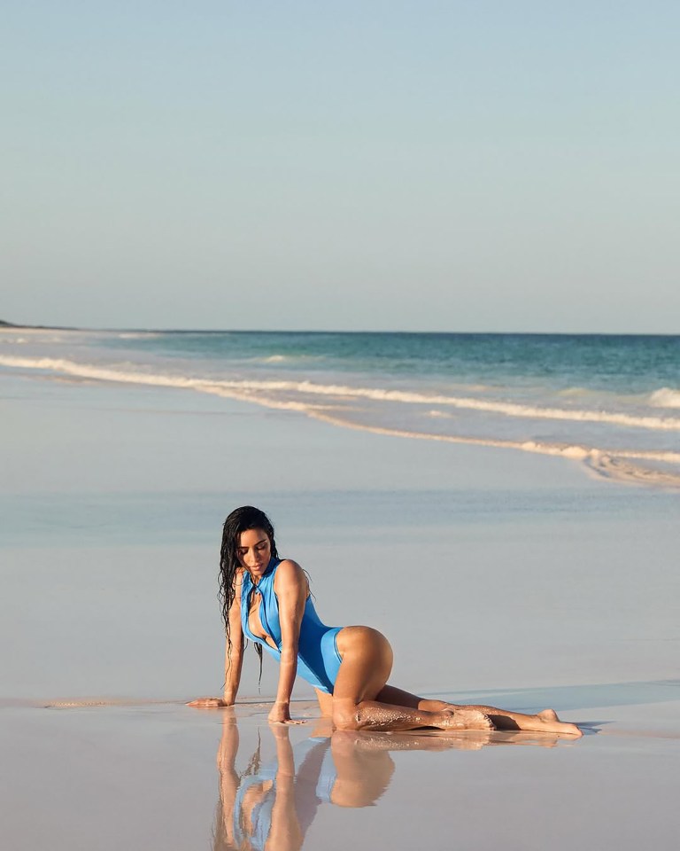 Woman in blue swimsuit kneeling on a beach.