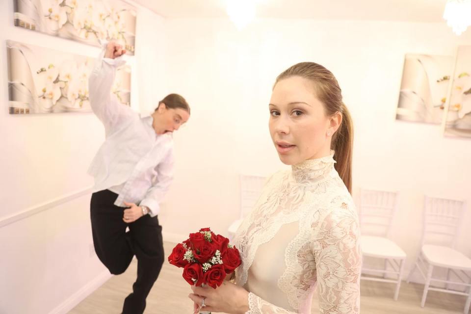 Woman in a lace wedding dress holding a bouquet, with a man in the background making a funny face.