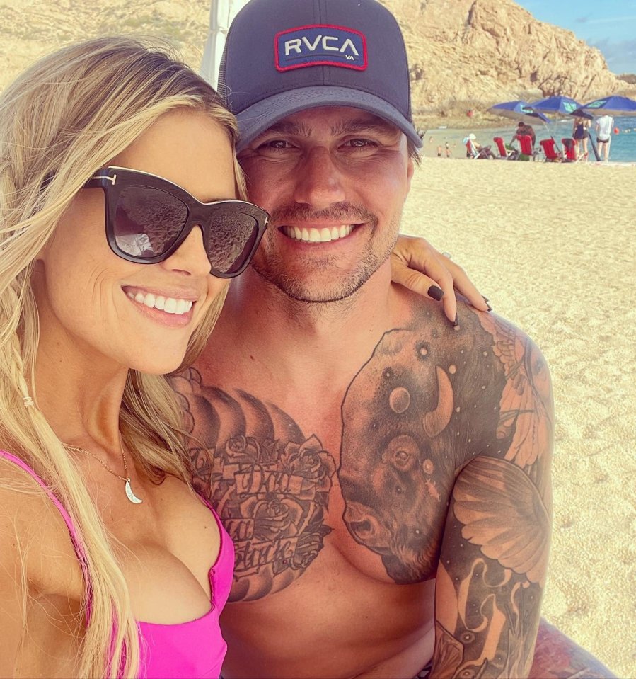 Christina Haack and Josh Hall embracing on a beach; she wears a pink bikini top and sunglasses, he has tattoos on his chest and arms.