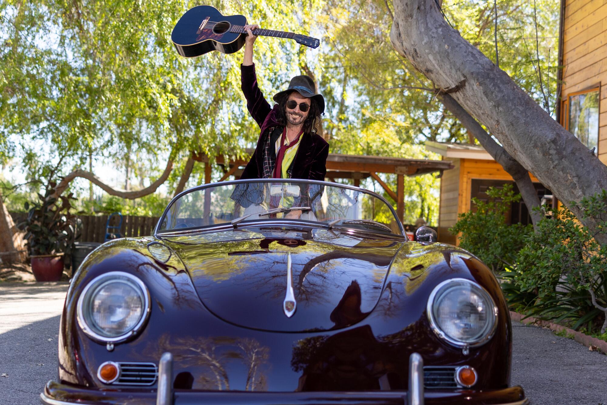 Mike Campbell sits up in his classic Porsche and holds his guitar in the air. 