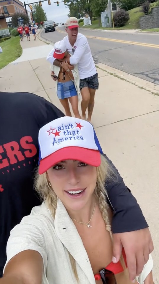 A woman wearing a "ain't that America" hat takes a selfie while a man carries another woman in the background.