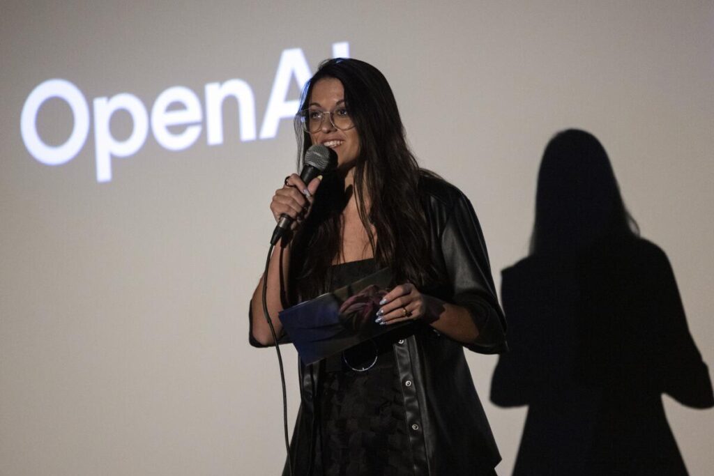 A woman with long hair, dressed in black, speaks into a microphone, the words "OpenAI" projected behind her