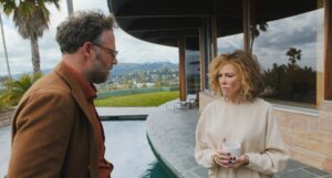 A man in brown suit looks at a woman in a white sweater holding a mug in her hands as they stand near a pool.