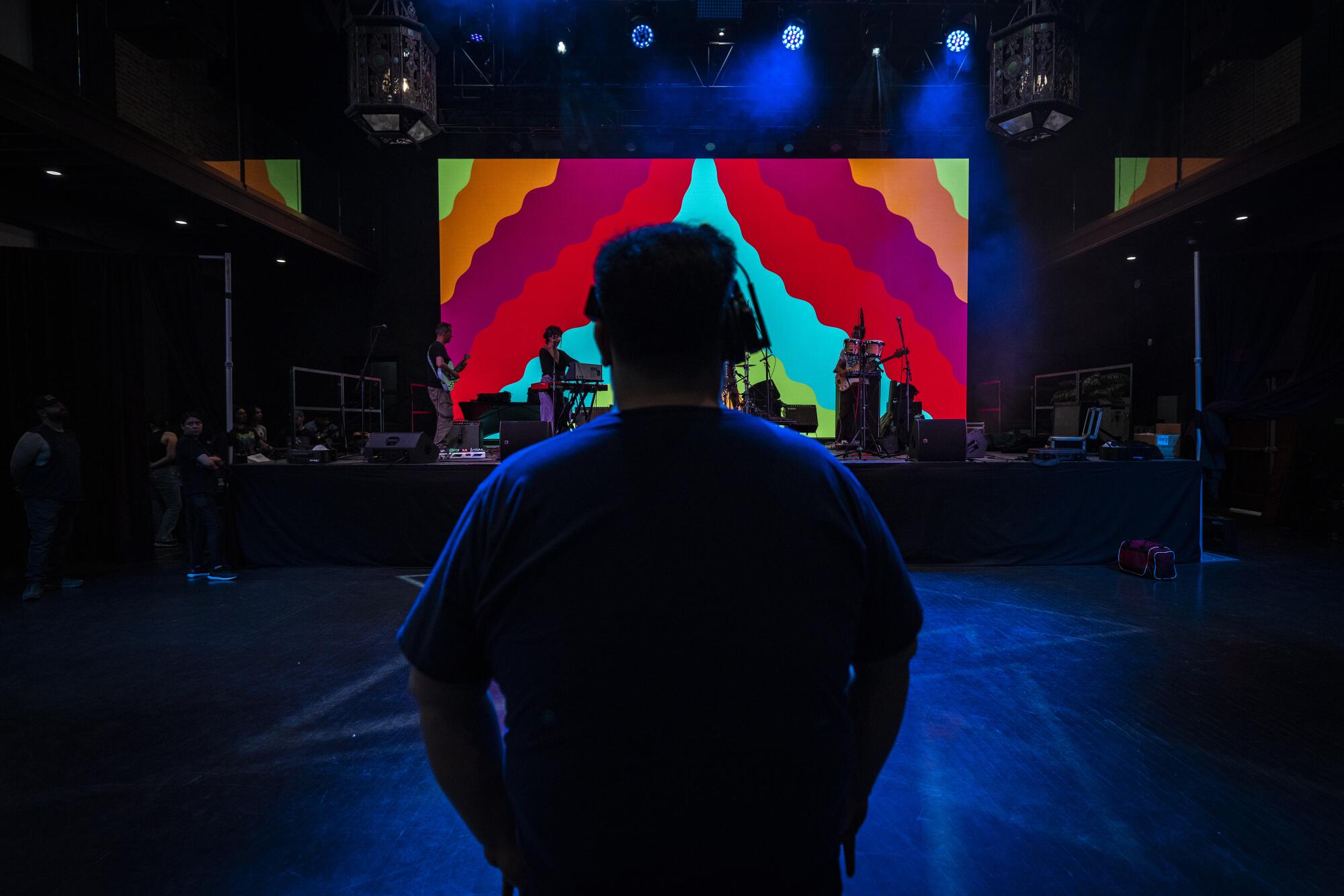 A sound technician wearing headphones, seen from behind, watches musicians perform onstage