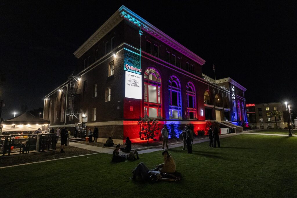 Concertgoers sit on break on the lawn in front of a building whose facade is lighted up with colored lights