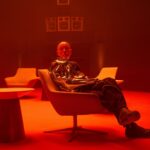 A man in a metallic coat and black trousers sitting in a chair next to a table in a room bathed in red light.