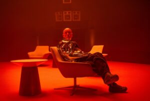 A man in a metallic coat and black trousers sitting in a chair next to a table in a room bathed in red light.