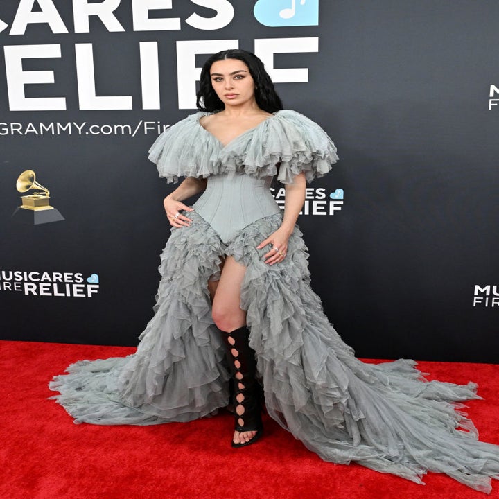 Person in an elaborate gown with ruffled, flowing layers and distinct shoulder design on a red carpet, promoting a music charity event