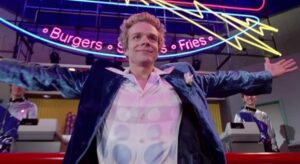 Man in a blue jacket with arms outstretched in a burger restaurant.