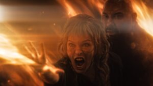 Milla Jovovich screams while using witch powers while Dave Bautista looks on in In the Lost Lands. They’re both surrounded by fire.