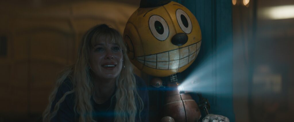 Millie Bobby Brown with long blonde hair next to a bobble-headed robot with a video projector in their chest in The Electric State.