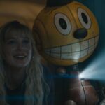 Millie Bobby Brown with long blonde hair next to a bobble-headed robot with a video projector in their chest in The Electric State.