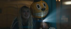 Millie Bobby Brown with long blonde hair next to a bobble-headed robot with a video projector in their chest in The Electric State.