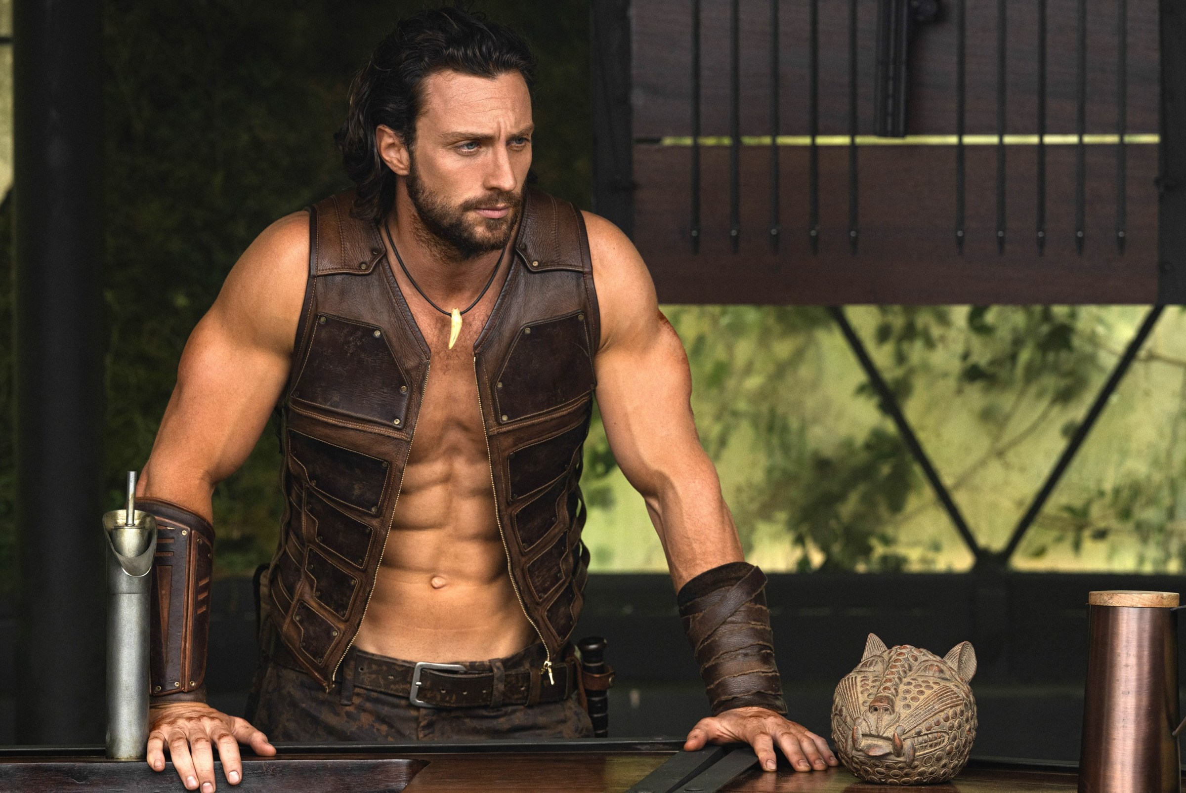 Kraven (Aaron Taylor-Johnson) stands with his hands on a railing, wearing a sleeveless, open leather vest and showing off more abs than seems humanly possible in Kraven the Hunter