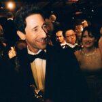 Adrien Brody, in a tuxedo, smiling, holding his Oscar. In the background are smiling onlookers.