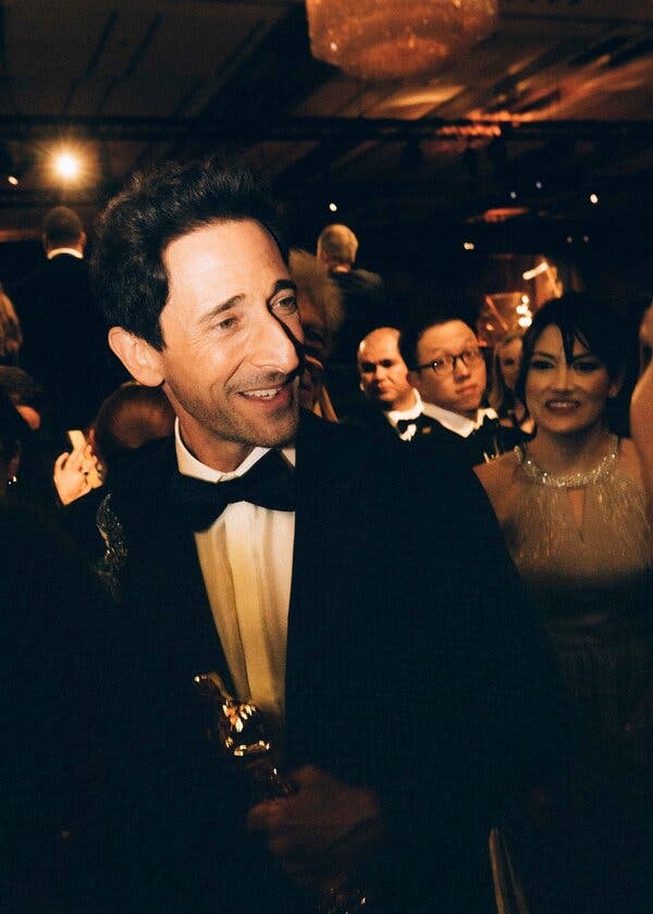 Adrien Brody, in a tuxedo, smiling, holding his Oscar. In the background are smiling onlookers.