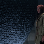 A woman in a green coat and orange cap standing in front of a massive spherical object studded with granite-like indentations in Tales from the Loop.