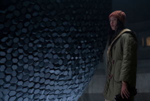 A woman in a green coat and orange cap standing in front of a massive spherical object studded with granite-like indentations in Tales from the Loop.