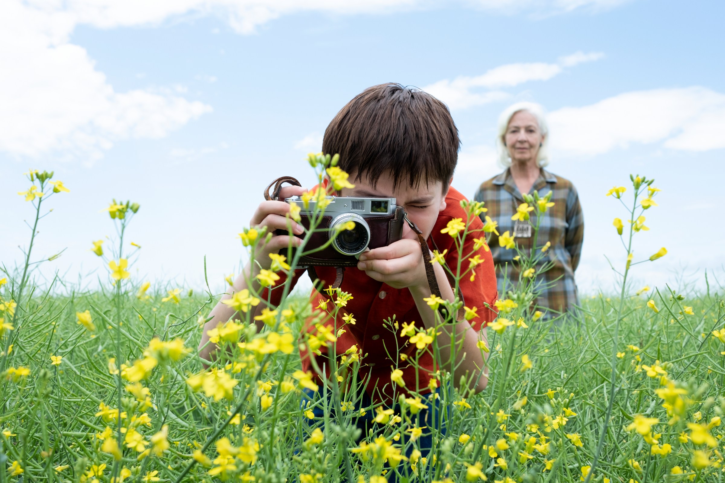 A boy in a bright red shirt uses an old-fashioned manual camera to take a closeup picture of a bright yellow flower as an older white-haired woman stands behind him and watches.